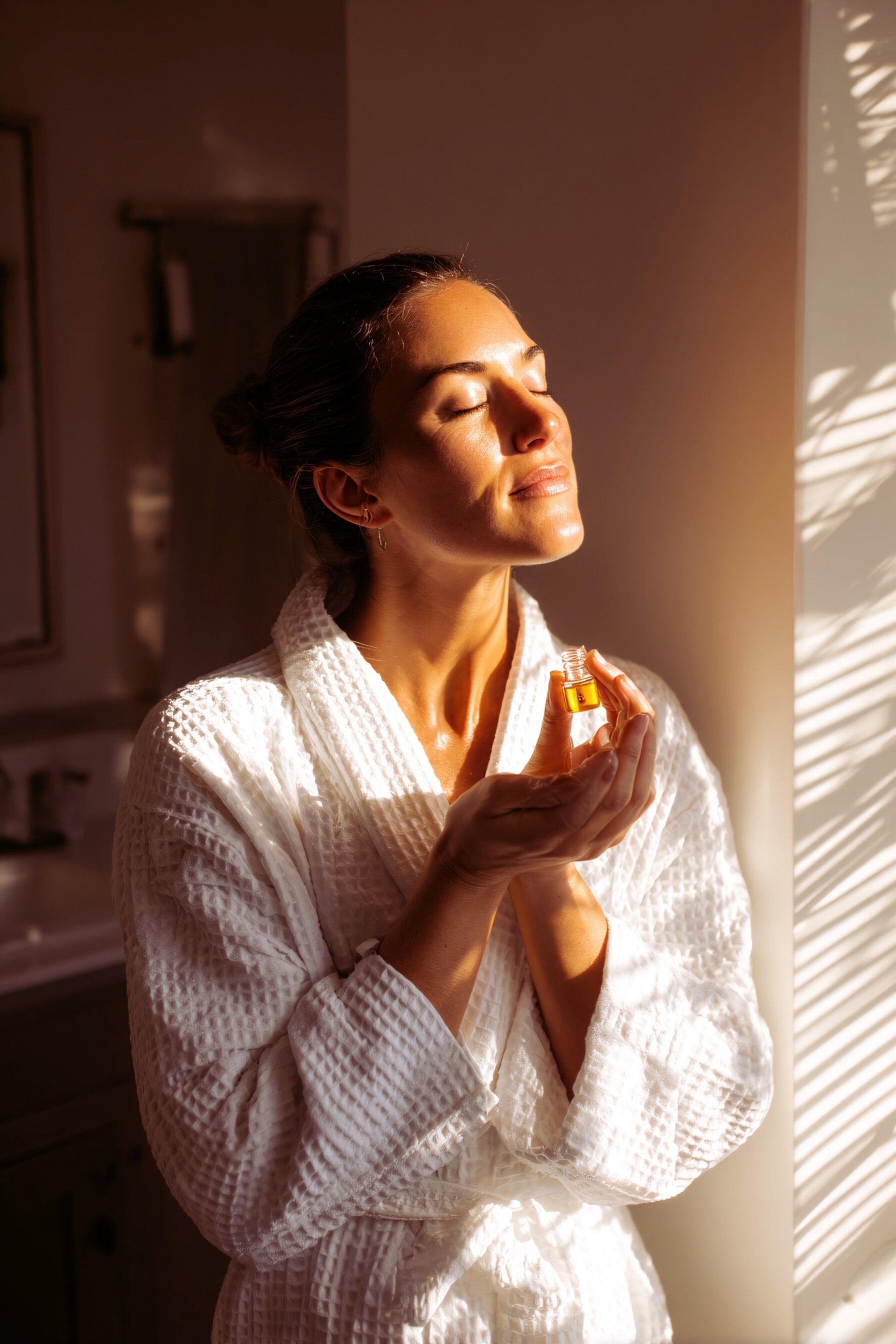 Woman in a white robe holding a bottle of perfume in a softly lit room.